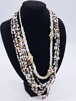 Front view of the full necklace displayed on a black bust stand, showing multiple strands of white, black, brown, gold, and pearl-like beads.