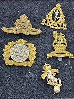 Five vintage military badges displayed on a dark fabric background showing various corps insignias including artillery and provost corps badges.
