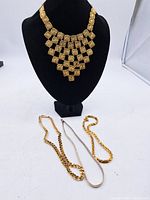 Four vintage costume jewellery necklaces displayed on and beside a black necklace bust. Includes a large gold-tone square patterned bib necklace, two medium length chains, and one small chain.