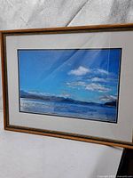 Framed pastel painting showing a scenic landscape with water, clouds, and mountains in the background, under glass in a wooden frame.