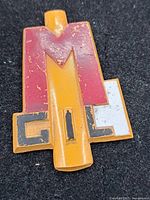 Front view of the Bakelite Youth Cap Badge showing the red, white, and yellow-orange colors and stylized MGIL design, with visible signs of wear and paint chipping.