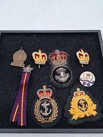 Top view of assortment of vintage British royal and naval badges including embroidered anchor patches, crown enamel pins, multicolored ribbon badge, and 1977 commemorative pin.
