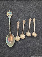 Full view of the five silver spoons on black fabric background showing the Alaska souvenir spoon and four smaller spoons.