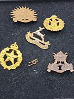 Photo showing five military badges laid out on black fabric, including a bronze St. Laurence Fusiliers badge with crossed rifles and maple leaf, gold-tone badges with crowned star and crowned stag, and other insignia.