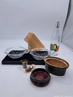 Lot of kitchen items including wooden cutting utensil block, two glass bowls, a glass bottle with floral decoration, a small metal cup, two cork stoppers, and a brown ceramic dish on a black tray.
