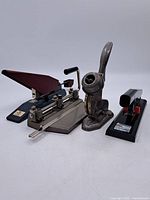Lot photo showing all four items: heavy-duty hole punch, two staplers including maroon one, and embossing seal press.