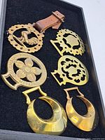 Overview of six vintage brass horse brasses displayed on black felt board, showing various designs and one mounted on leather strap.