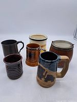 Six vintage and antique drinkware items including pottery mugs, a pewter cup, and a wooden barrel-style mug displayed on a white surface.