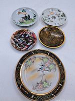 Five vintage decorative dishes arranged on a white surface showing different artistic styles including cloisonne and painted ceramic.