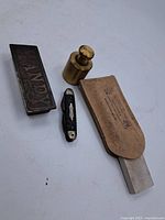 Vintage items displayed on white surface: Arkansas stone in metal box labeled HANDY, brass weight, small pocket knife, leather sharpener stone.