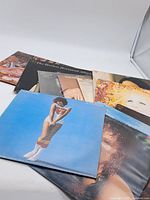 Lot of seven vintage vinyl records placed overlapping on a white surface. One album cover shows a woman wearing a Superman t-shirt against a blue background.
