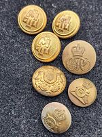 Seven military buttons on black fabric: 4 Italian buttons with 'M' and crown, plus 3 British buttons with crown, cannon, and coat of arms.
