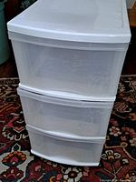 Front view of the three-drawer white plastic storage unit on a patterned rug, showing its used condition and drawer fronts.