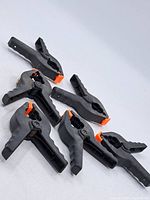 Six black nylon spring clamps with orange gripping tips arranged on a white surface.