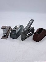Four vintage office items including two staplers and two tape dispensers laid in a row on white background