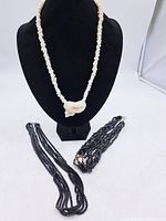 Three costume jewellery necklaces displayed on black stands: long white bead necklace with shell-like pendant, multi-strand black bead necklace, and a short white bead necklace.