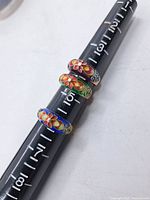 Three vintage enameled silver rings stacked on a ring sizing mandrel, showing colorful floral enamel designs and size markings on the mandrel.