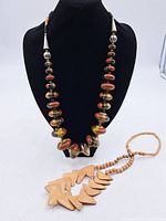 Photo of two necklaces displayed on a black jewelry bust: one with large multicolored glass beads and metal separators and the other wooden with carved pendant pieces.