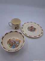 Photo showing a Bunnykins mug, shallow bowl, and plate with bunny motifs around rim and Bunnykins scenes in center.