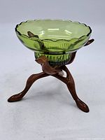 Side angle showing the green glass bowl with scalloped edges resting on a carved wooden stand with bird features on the legs.