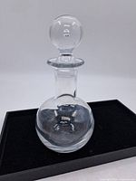 Full view of the entire glass decanter including the round glass stopper on top, placed on a black base.