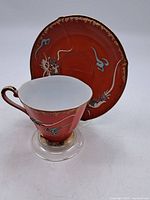 Front view of teacup positioned in front of saucer showing reddish-orange color and blue dragon design with gold trim.