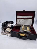 Photo showing the vintage black rotary phone with coiled cord and the Monroe adding calculator set inside a black carrying case with burgundy interior and attached lumber tables.