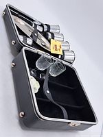 Photo of opened black leatherette Trav-L-Bar case showing six stainless steel cups with leather securing strap and two etched glass stemmed glasses with leather strap.