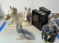 Photo showing brass horse head bookends, black monogrammed bookends, glass dolphin figurine, and colorful glass decanter