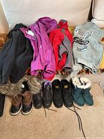 Photo shows five pairs of women's boots (brown fur-trimmed winter boots, black Cat work boots, black Brahma work boots, teal fur-trimmed winter boots, brown fur-lined winter boots) lined up on floor with three jackets (black, purple raincoat, red and gray fleece) and gray Speedster sweatshirt with motorcycle print on a couch.