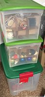Two stacked plastic storage boxes with green lids and a tall clear container with a red latch; inside the containers, various Christmas ornaments are visible including several baubles and shaped ornaments.