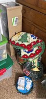 Christmas tree skirt with holiday needlepoint design, partially inside a green bucket, various boxes and merchandise around it