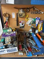 Wide view of the pegboard displaying a variety of hand tools, fasteners, sockets, and caulking applicators arranged on and in front of pegboard. Visible are hammers, two caulking guns, packaged nails and screws, roll of tape, and grout saw.