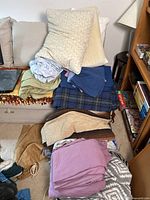 Photo showing stacked linens and blankets on a couch, including pillows, sheets, and a plaid comforter duvet.