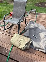 Outdoor rocking chair on wood deck with curved metal rockers and gray textured fabric seat and backrest, accompanied by two folded covers: one gray lounge chair cover and one light green fitted chair cover.
