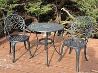 Wide shot of black wrought iron bistro-style table and two matching chairs on wooden deck outdoors.
