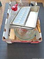 Photo of Pyrex mixing bowls nested at bottom, metal baking trays, plastic ice cube trays, wooden rolling pins, glass bottles, and other kitchen tools arranged in box