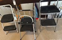 Front view of two single step stools and two double step stools with metal frames and black plastic textured steps, on carpeted floor.