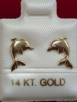 Pair of 14KT gold dolphin stud earrings displayed on white textured holder with '14 KT GOLD' printed in gold lettering.