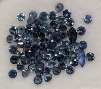 Close up of multiple loose blue sapphire gemstones on a light surface, showing round and some marquise shapes