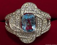Close-up photo of a silver ring with a cushion-cut blue topaz center stone surrounded by small clear accent stones set in an ornate looped pattern.