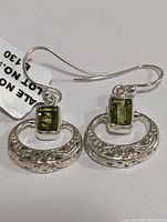 Front view showing pair of silver earrings with green rectangular peridots and textured crescent decoration.