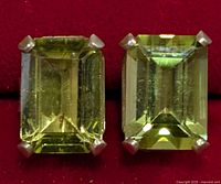Close-up photo showing two rectangular emerald-cut peridot stones set in silver prongs on a red background.