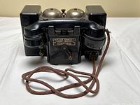 Front view showing handset on cradle, dual metal bells on top, crank lever and connection posts on front, cloth-covered cord coiled in front.