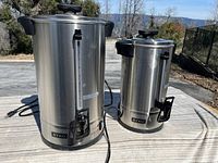 Two stainless steel Sybo coffee or beverage urns of different sizes shown side by side on a table outdoors, power cords visible.