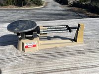 Photo showing full side view of Ohaus Triple Beam Balance scale with visible weighing platform and sliding weights on beams on pavement outdoors.