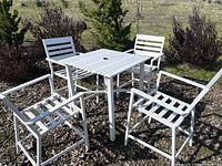 Image showing the entire set of white aluminum outdoor furniture consisting of one square table and four chairs arranged outdoors on a stone surface with plants in background.