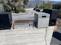 View of stereo amplifiers, Onkyo tuner, and three black subwoofers on flat surface outdoors.