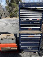 A wide view showing all three tool boxes placed outside on asphalt: the largest blue Craftsman rolling tool chest with stickers, a mid-sized black plastic box with metal latch locks, and a smaller red metal tool box with drawers.