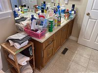 Bathroom counter with numerous personal care and cleaning products, plastic storage baskets, and wooden cabinetry underneath. Small woven shelf at the side with folded towels.